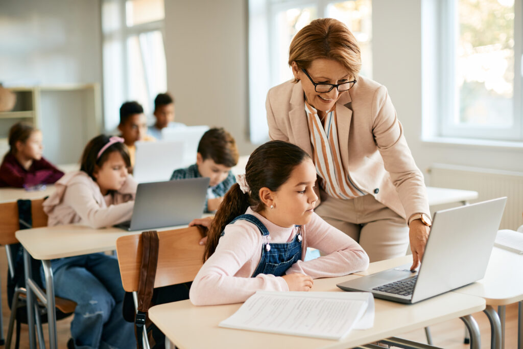 Happy teacher assisting schoolgirl in using laptop during computer class in the classroom.